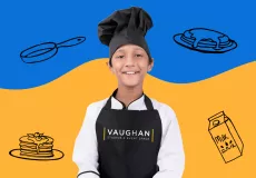 A young male kid chef somewhere between the ages of 7 and 9 smiling straight at the camera wearing a black baker's cap, and what appears to be the traditional chef's whites including a white long-sleeved jacket with black trim, and a black apron with the Vaughan Studios & Event Space logo on the front. 