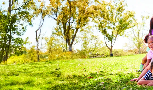 A grass field with trees in the background with kids huddled on top of each other