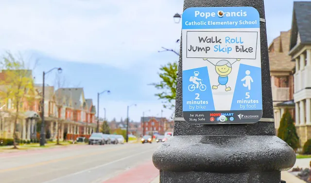 Sign for Pope Francis Catholic Elementary School promoting walking and biking, shown on a post along a residential street with houses and cars in the background