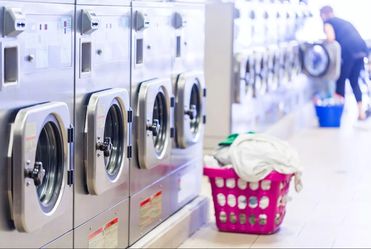 Laundromat with basket on the ground and person removing laundry from a dryer