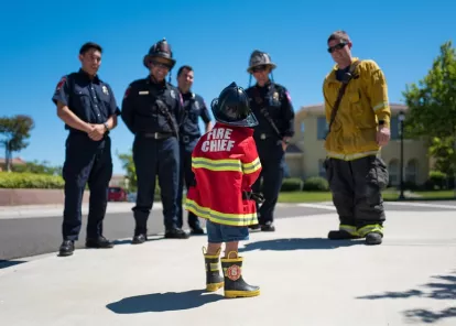 child in a fire chief jacket standing in a semi-circle of adult firefighters