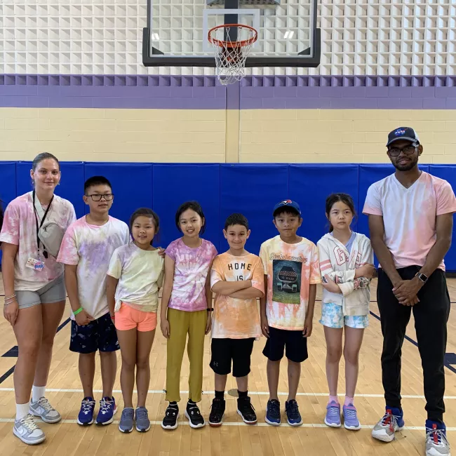 A group of summer campers and their camp counsellors posing in an indoor gymnasium with a basketball hoop in the background.