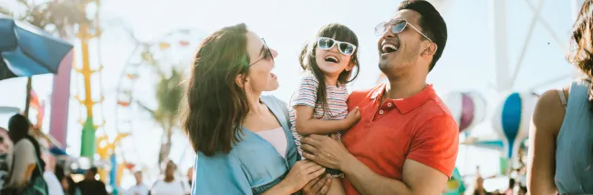 A mother and father holding their daughter while smiling while at an outdoor public event