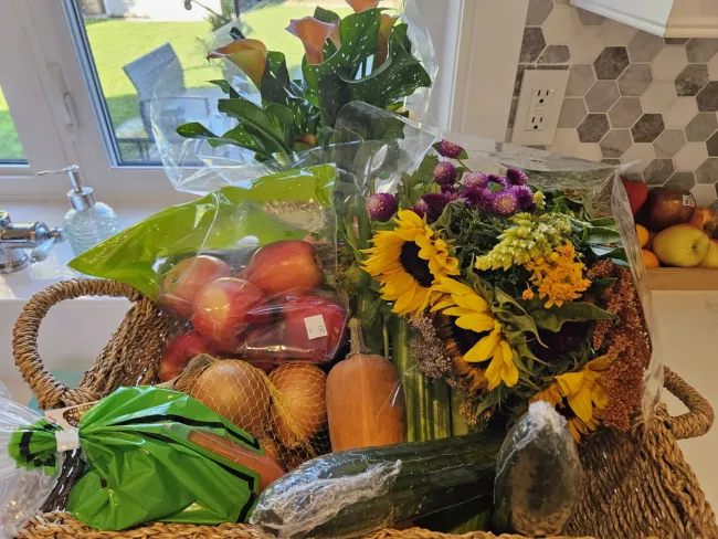 Woven basket on a kitchen counter with fresh produce - apples, onions, cucumber, butternut squash - and sunflowers, purple flowers, and a calla lily