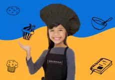 A young female kid chef somewhere between the ages of 7 and 9 smiling straight at the camera wearing a black baker's cap, a grey long-sleeved shirt, and a black apron with the Vaughan Studios & Event Space logo on the front.