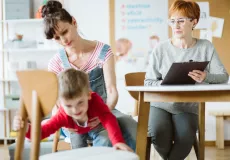A young mother with her squirming child sitting at a table with an older woman wearing glasses and holding a clipboard who appears to be a therapist or counsellor