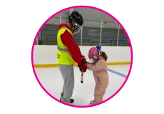 A skate instructor assisting a young skater on the ice in an indoor arena.