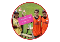 A group of three volunteers wearing bright orange or yellow vests holding a bright pink sign that says "Volunteer Area."