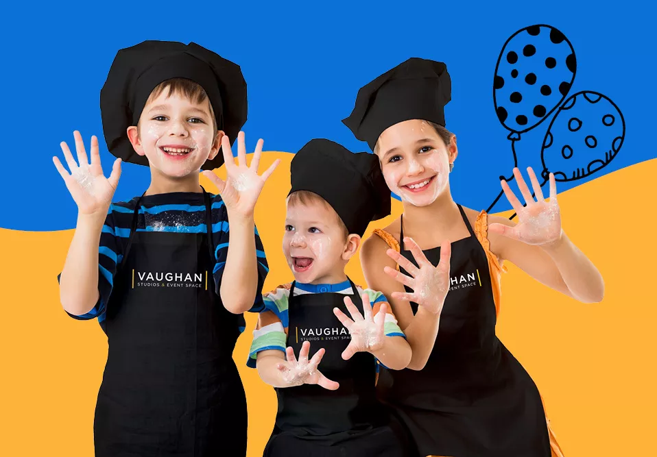Three young kid chefs between the ages of 5 and 12 smiling with both hands up and palms facing forward to show the flour on their hands, all three wearing a black baker's caps and black aprons with the Vaughan Studios & Event Space logo on the front. 