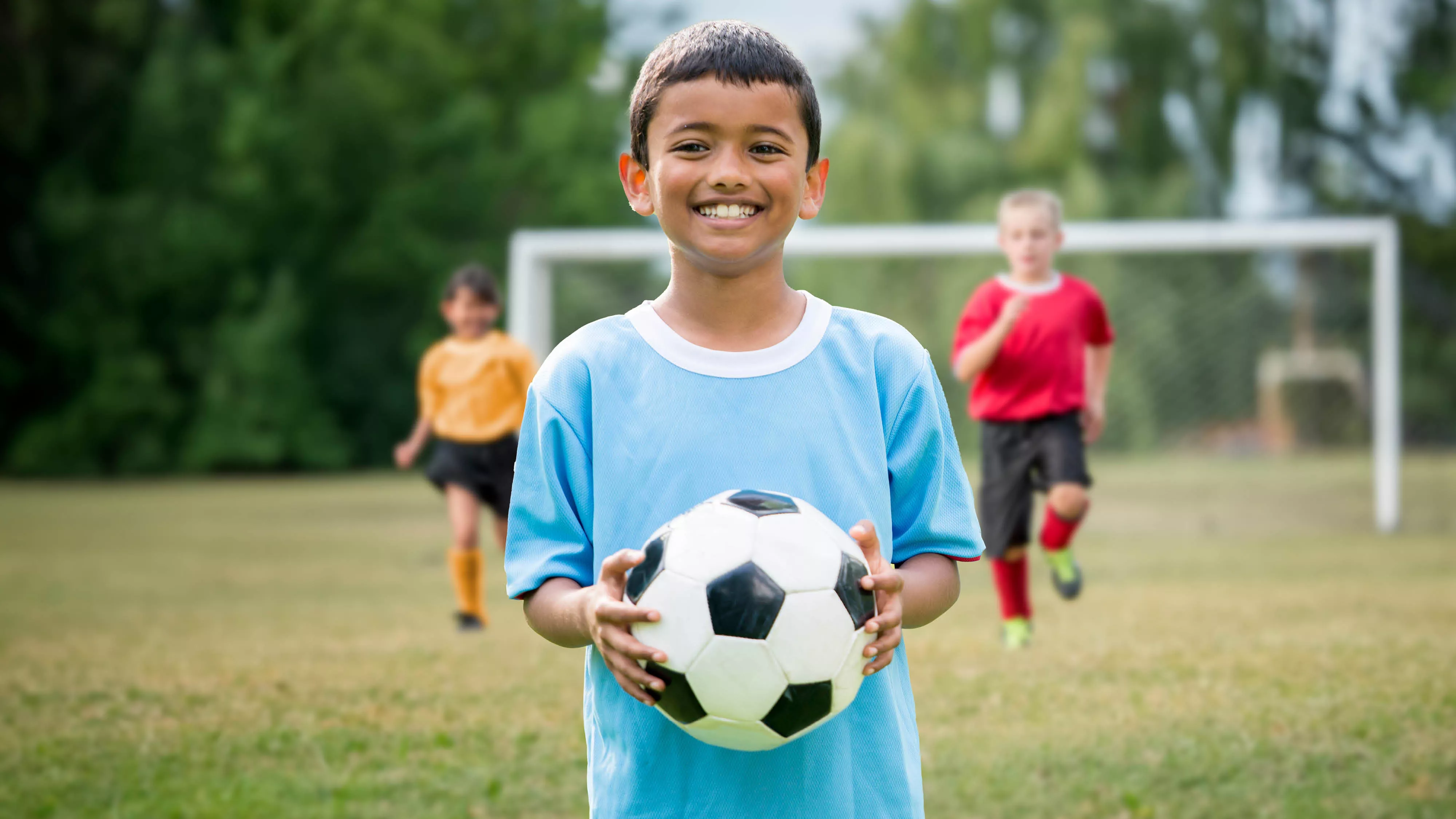 Little boy holding a soccer ball on the field with two kids running behind him