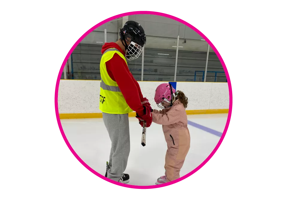 A skate instructor assisting a young skater on the ice in an indoor arena.