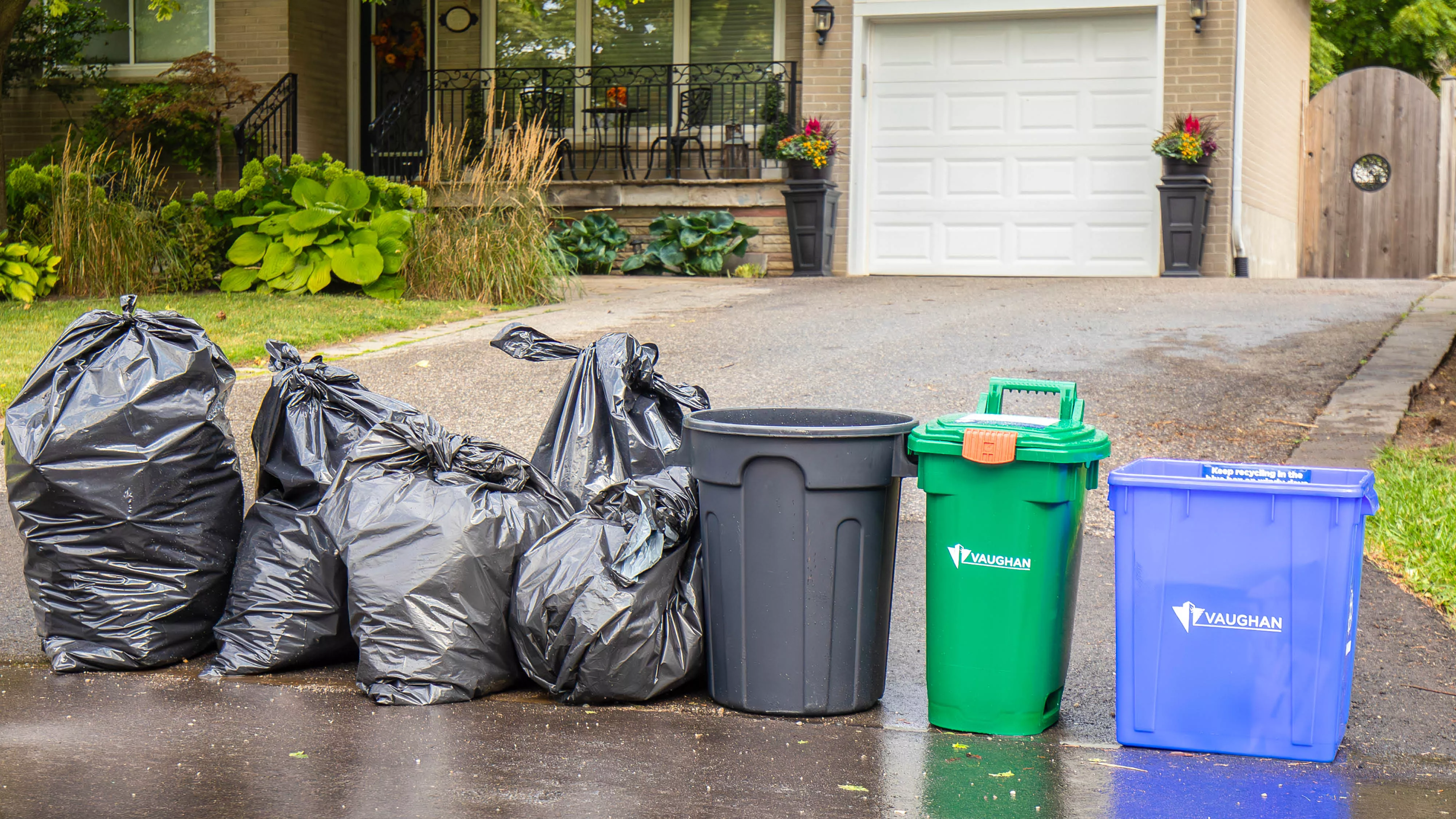 four garbage bags, one green bin and one blue box at the end of a driveway