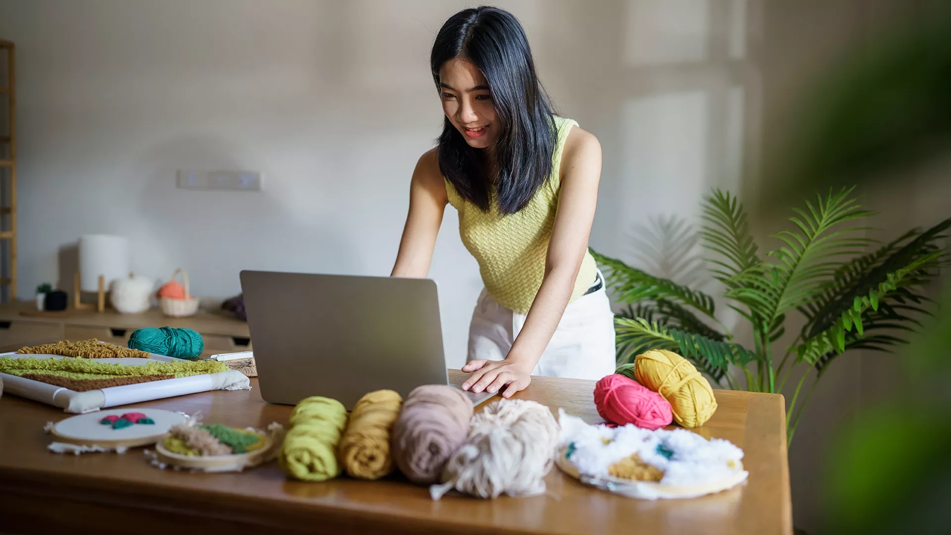 Woman standing at a desk looking at her laptop. Desk has balls of yarn on it. 