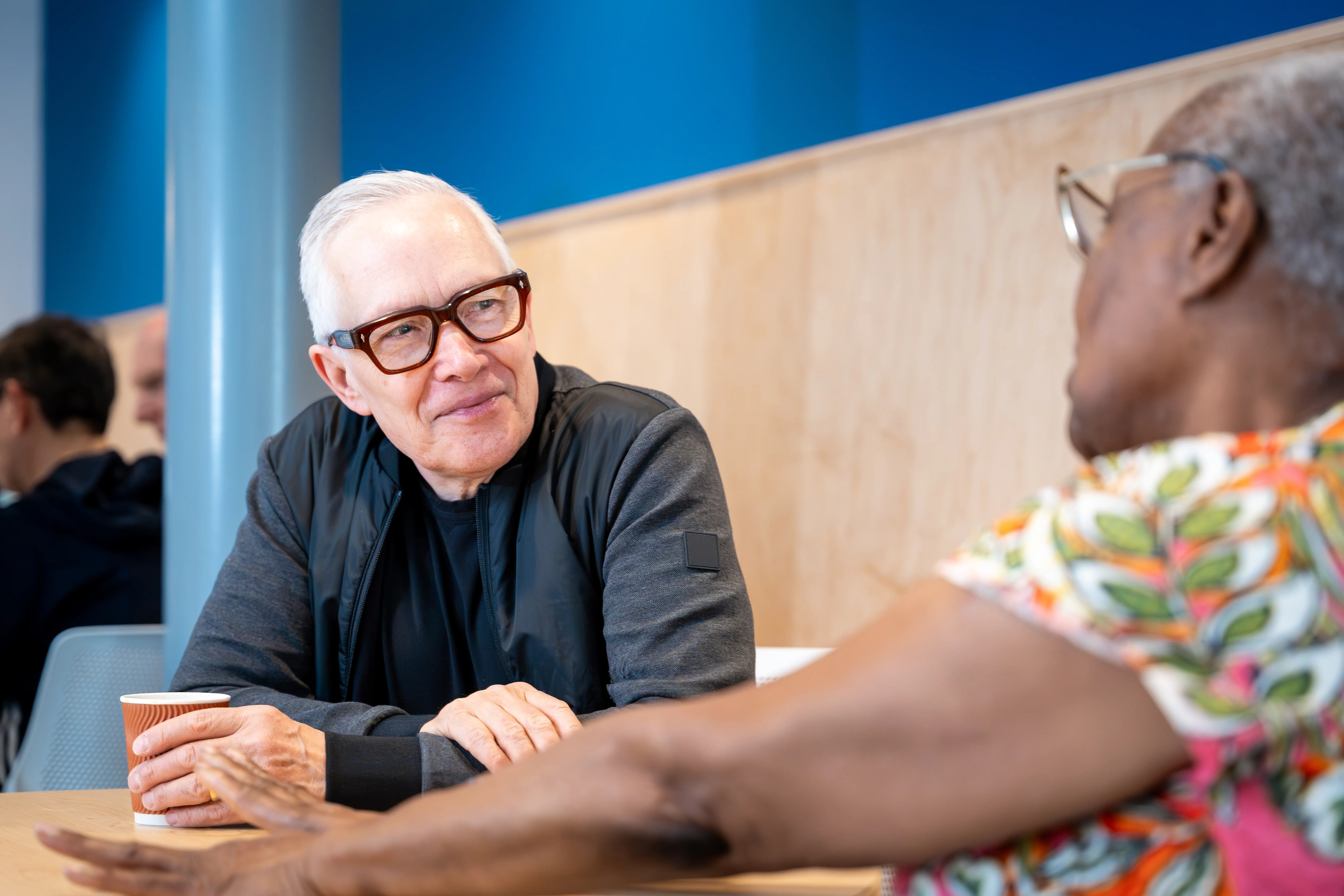 2 individuals talking. Man holding a cup of coffee